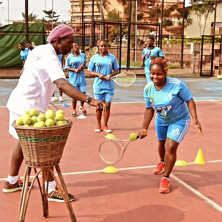A coach holding out a basket of tennis balls as young BRIGHT players train with their rackets on the court